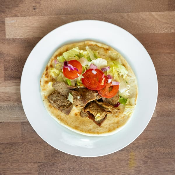 Open flatbread topped with sliced gyro meat, lettuce, tomatoes, and red onions on a white plate.