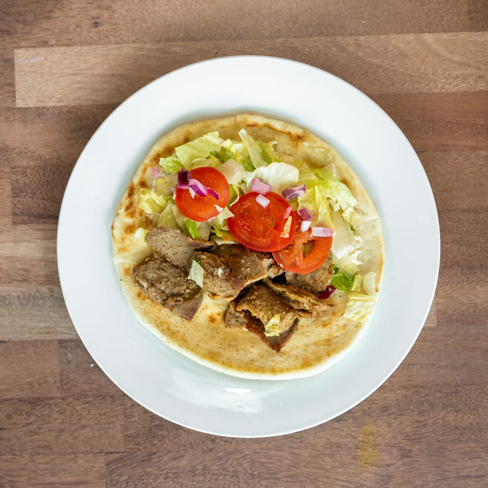 Open flatbread topped with sliced gyro meat, lettuce, tomatoes, and red onions on a white plate.