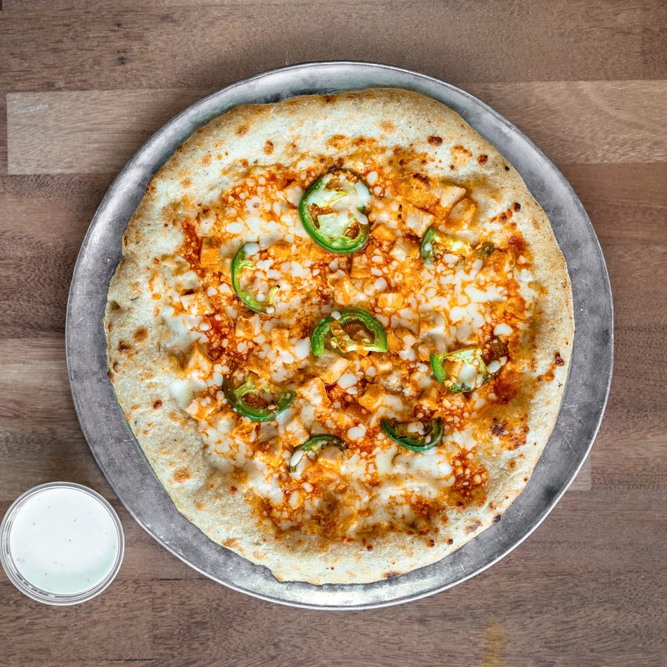 Cheesy pizza topped with diced chicken and sliced green jalapeños on a metal tray, accompanied by a cup of white dipping sauce.