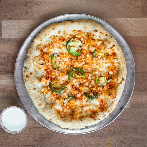 Cheesy chicken pizza topped with sliced jalapeños on a metal tray, accompanied by a cup of white dipping sauce.