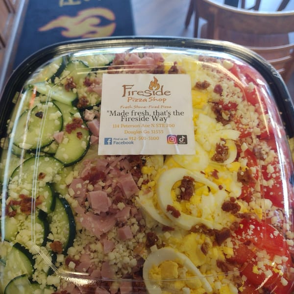 Fireside Pizza Shop salad platter with cucumbers, ham, crumbled cheese, eggs, tomatoes, and bacon in a takeout container.