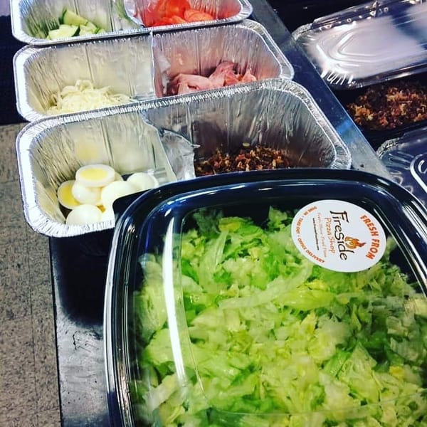 Fresh chopped salad bar ingredients in foil trays, including lettuce, cucumbers, tomatoes, onions, eggs, shrimp, and toppings.