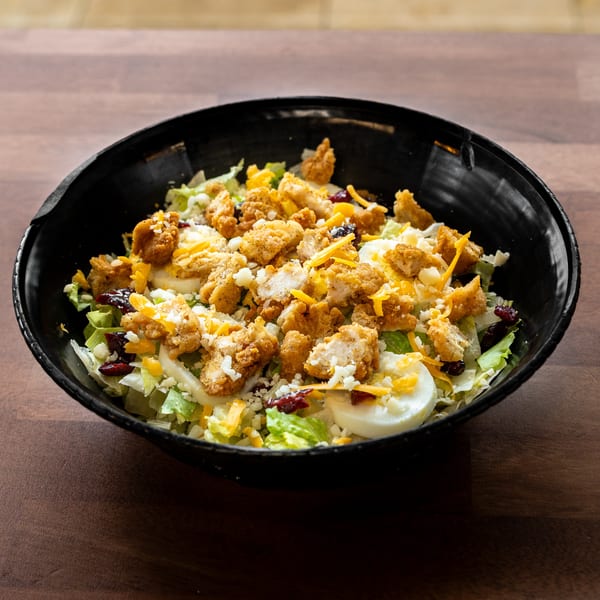 Black bowl of lettuce salad topped with crispy chicken, sliced eggs, shredded cheese, cranberries, and dressing on a wooden table.